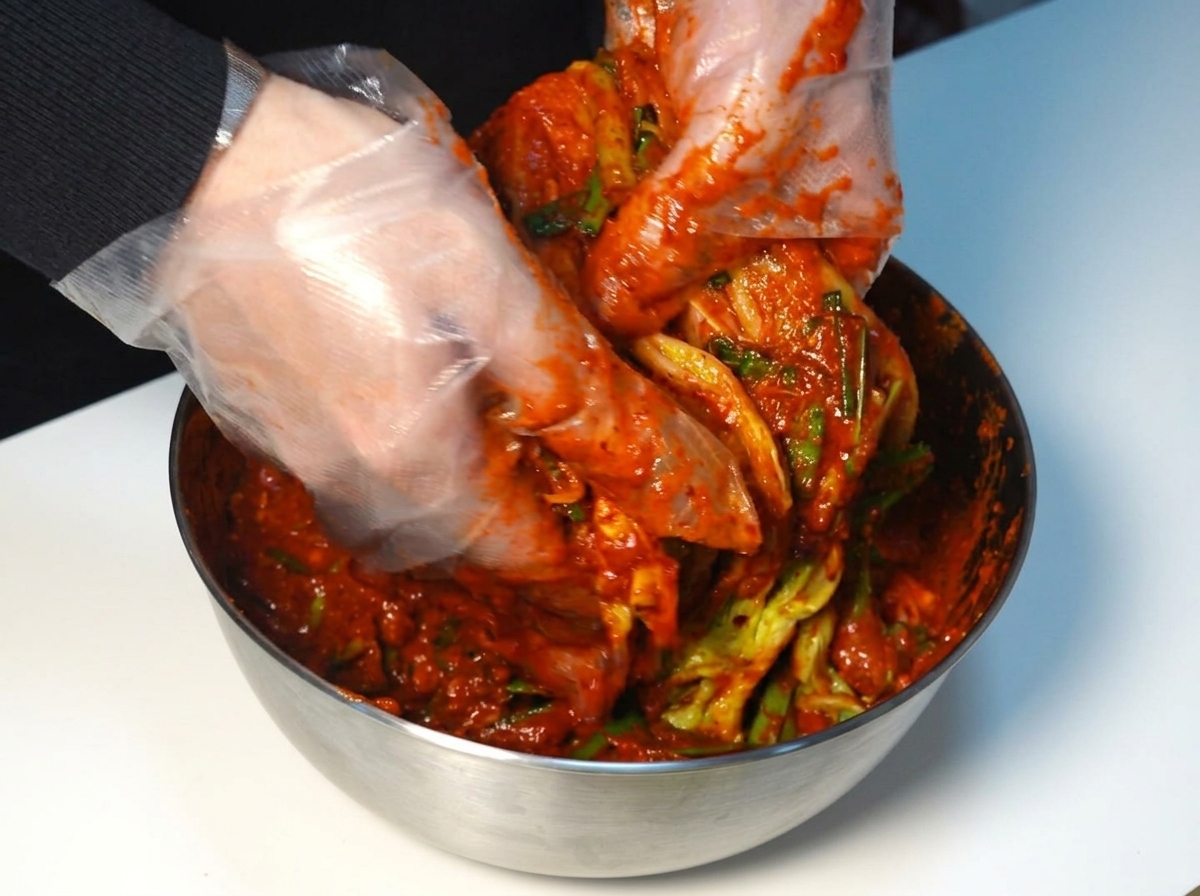 Ręce w rękawiczkach rozprowadzające czerwoną pastę kimchi na liściach kapusty.