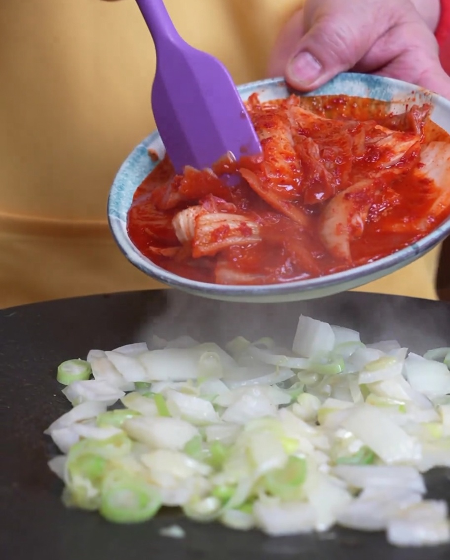 Kimchi dodawane do patelni z podsmażoną zieloną cebulką.