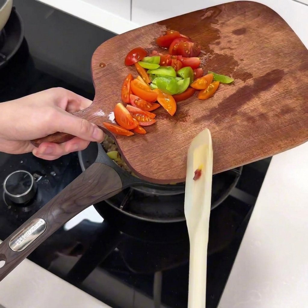 Świeżo pokrojone pomidorki koktajlowe i paski zielonej papryki przenoszone z drewnianej deski na patelnię.