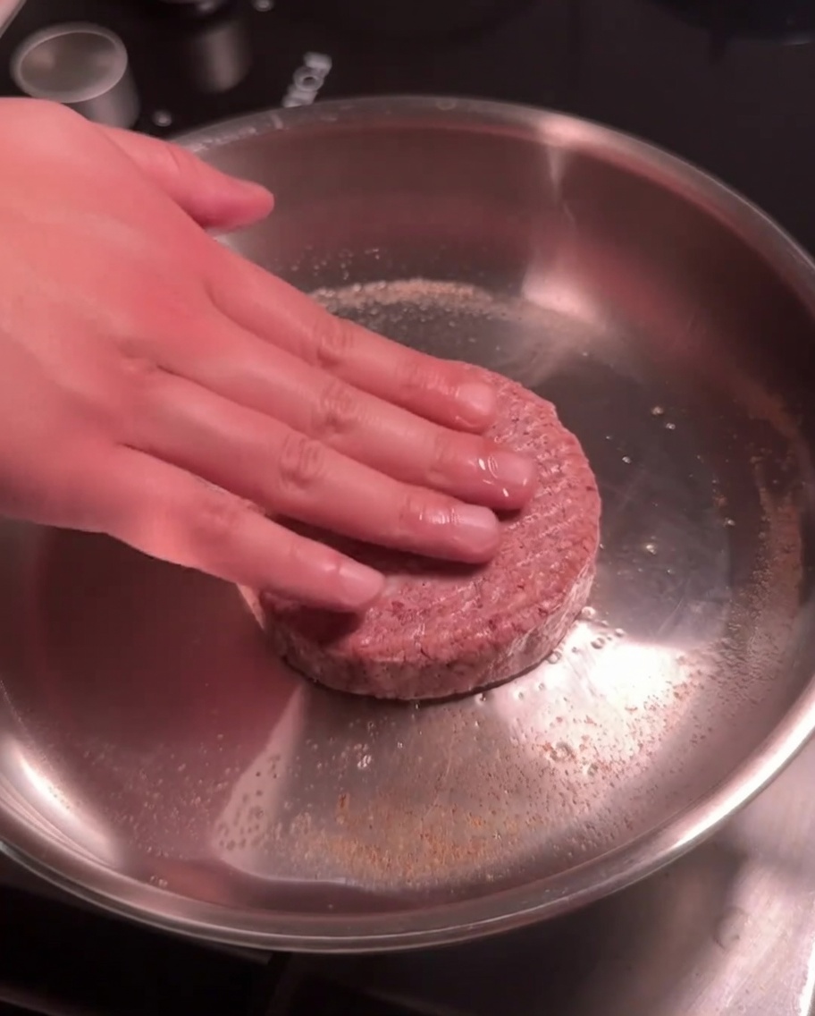 Surowy kotlet wołowy dociskany do gorącej patelni ze stali nierdzewnej podczas smażenia.