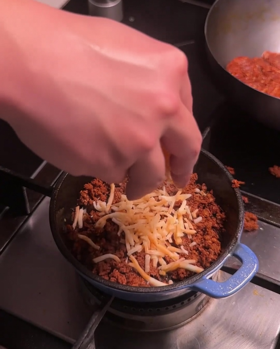 Tarty ser posypywany na usmażone mięso chorizo w małym niebieskim garnku na kuchence.