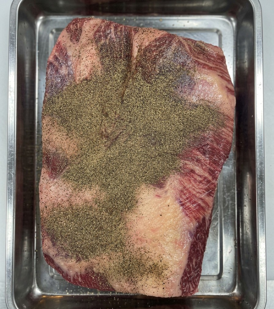 Dłonie w rękawiczkach wciskające czarny pieprz w powierzchnię mostka Wagyu w celu stworzenia równomiernej warstwy.