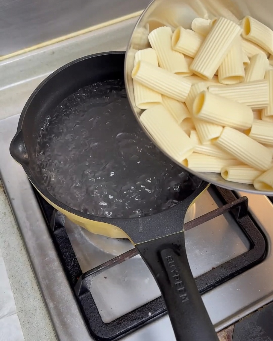 Namoczony makaron rigatoni wsypywany z metalowej miski do garnka z wrzącą wodą na kuchence gazowej.