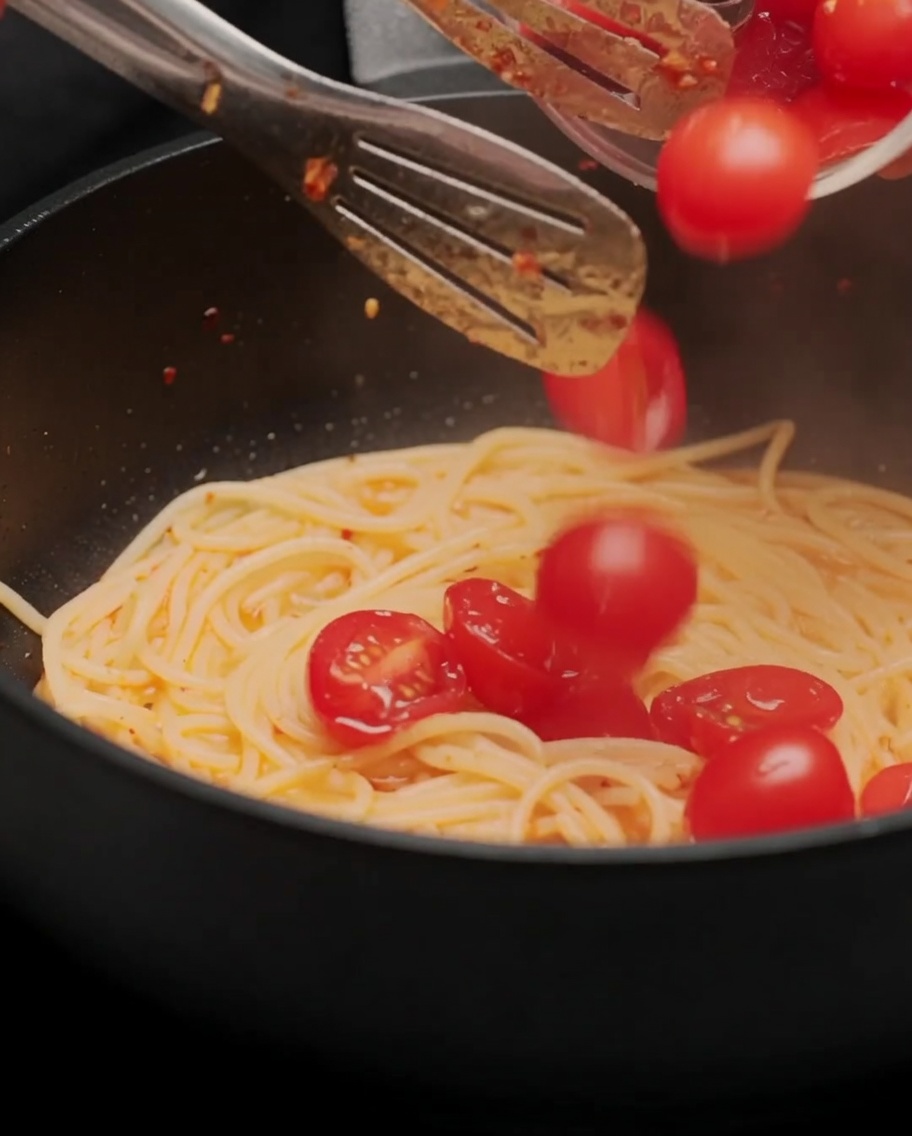 Przekrojone na pół pomidorki koktajlowe dodawane do makaronu na patelni.