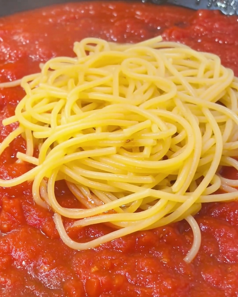 Łyżka skrobiowej wody z gotowania makaronu dodawana do spaghetti z sosem na patelni.