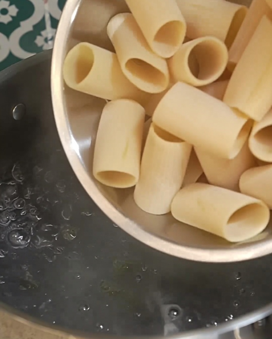 Surowy makaron rigatoni wsypywany do garnka z wrzącą wodą.