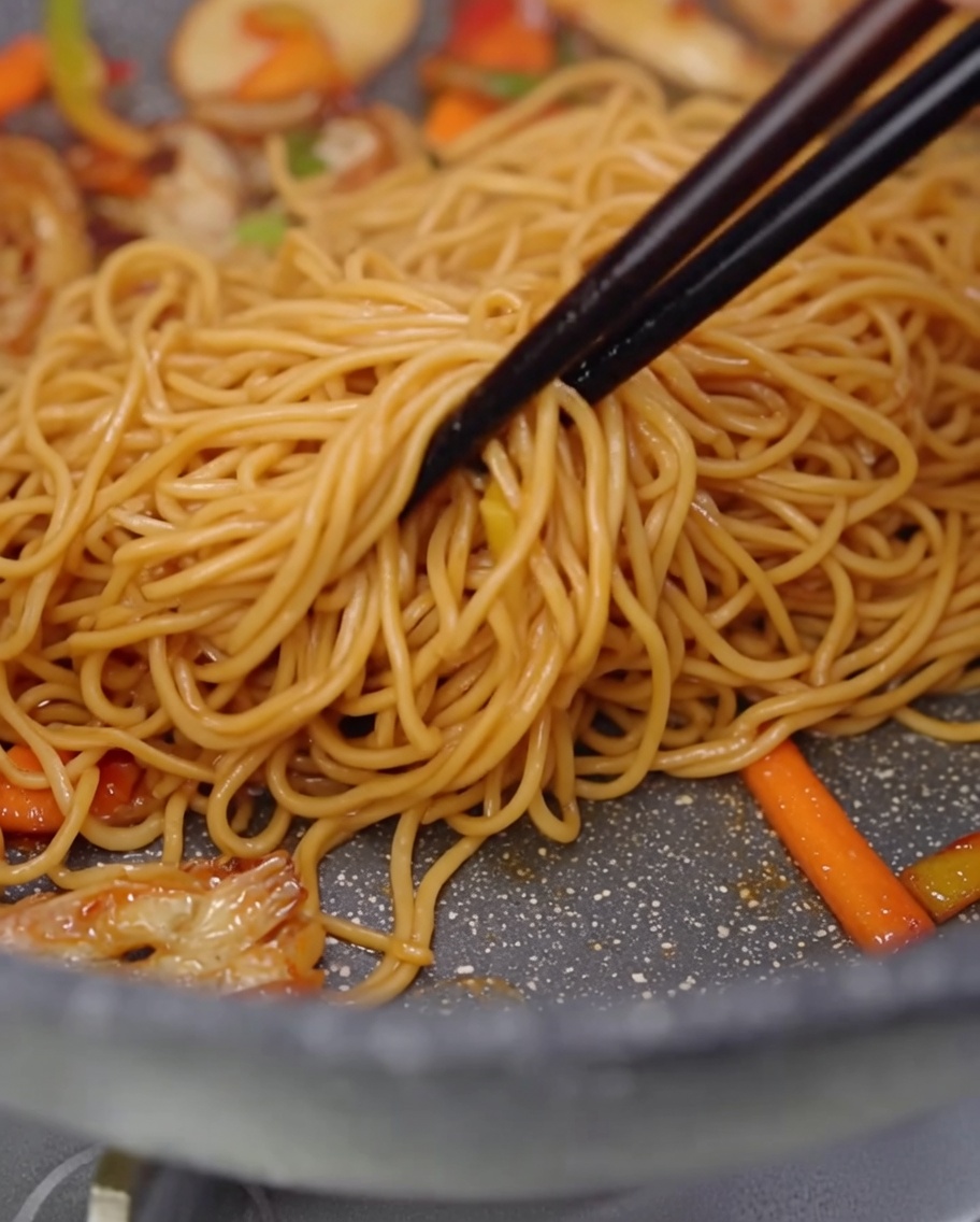 Spaghetti, krojone grzyby i papryka mieszane na patelni z użyciem drewnianych pałeczek.