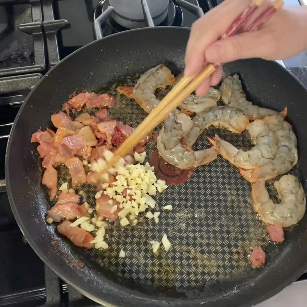 Drobno posiekany czosnek rozsypywany na krewetki i bekon na patelni.