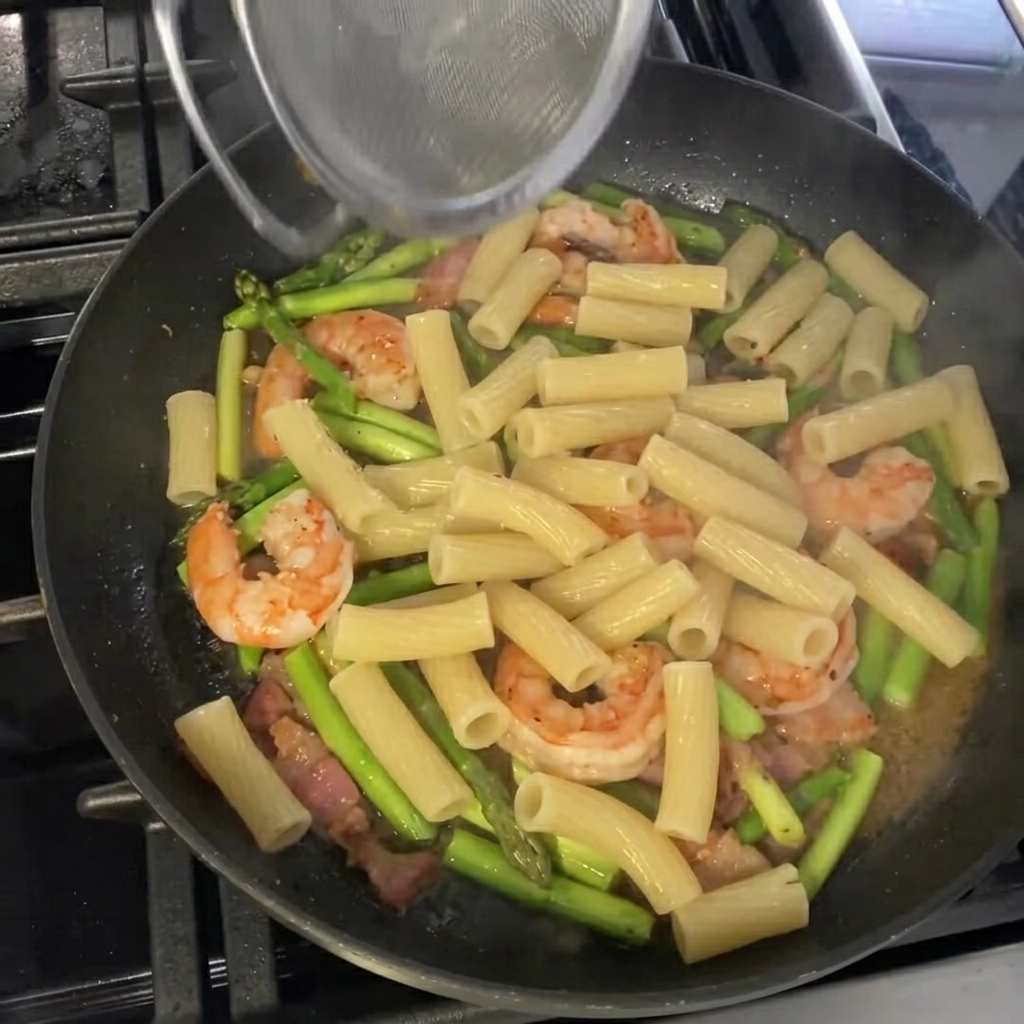 Ugotowane rigatoni wrzucane na patelnię ze szparagami i krewetkami, gotowe do finalnego doprawienia.