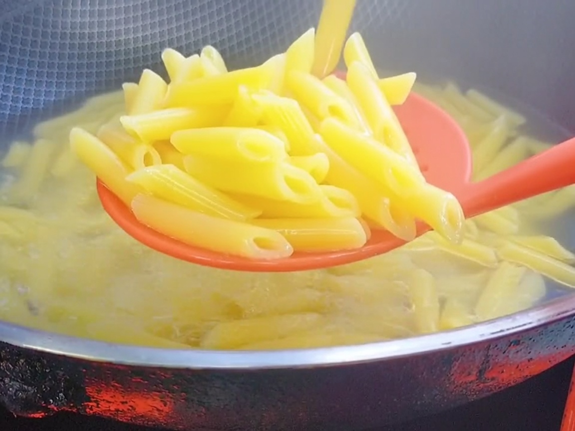 Czerwona silikonowa łyżka cedzakowa wyjmująca ugotowany makaron penne z wrzątku.