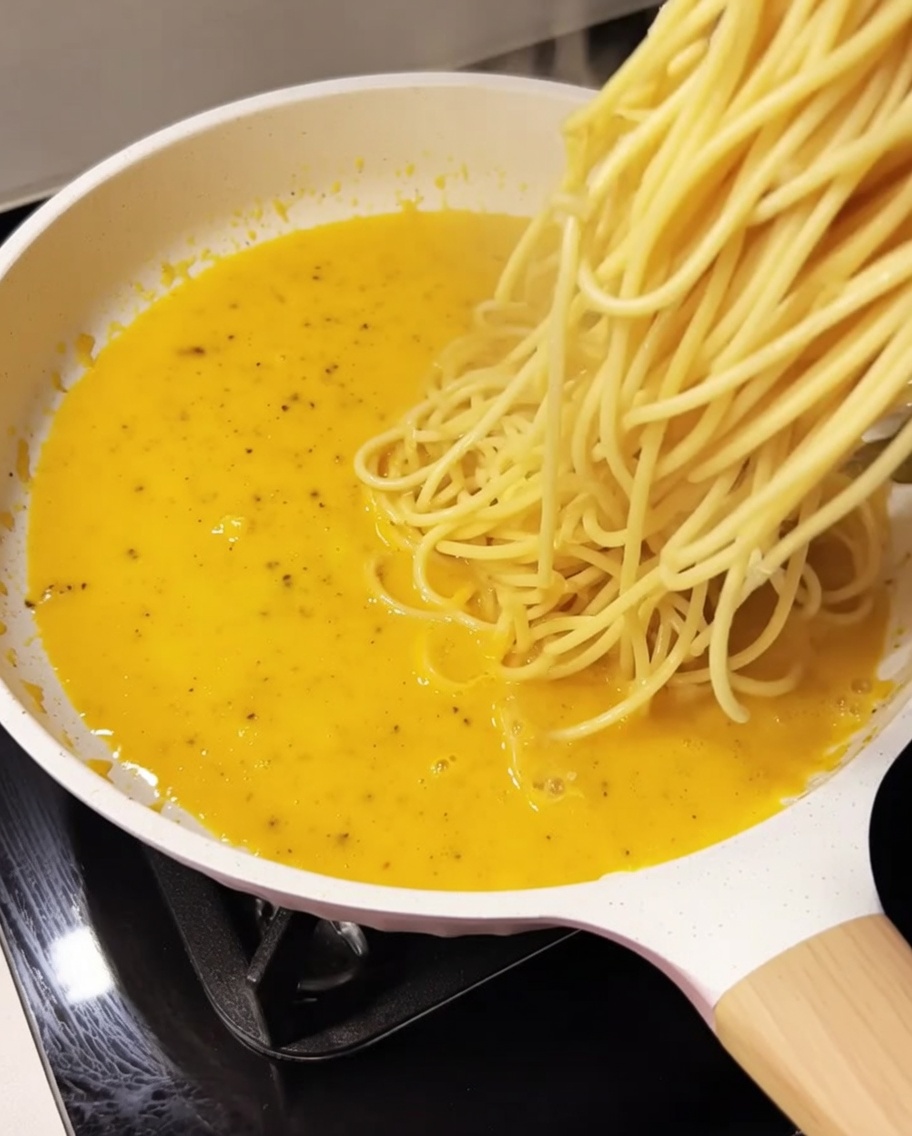 Ugotowane spaghetti dodawane do kremowego, żółtego sosu na białej patelni.