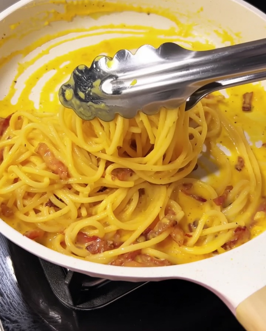 Szczypce mieszające spaghetti z sosem i guanciale na patelni.