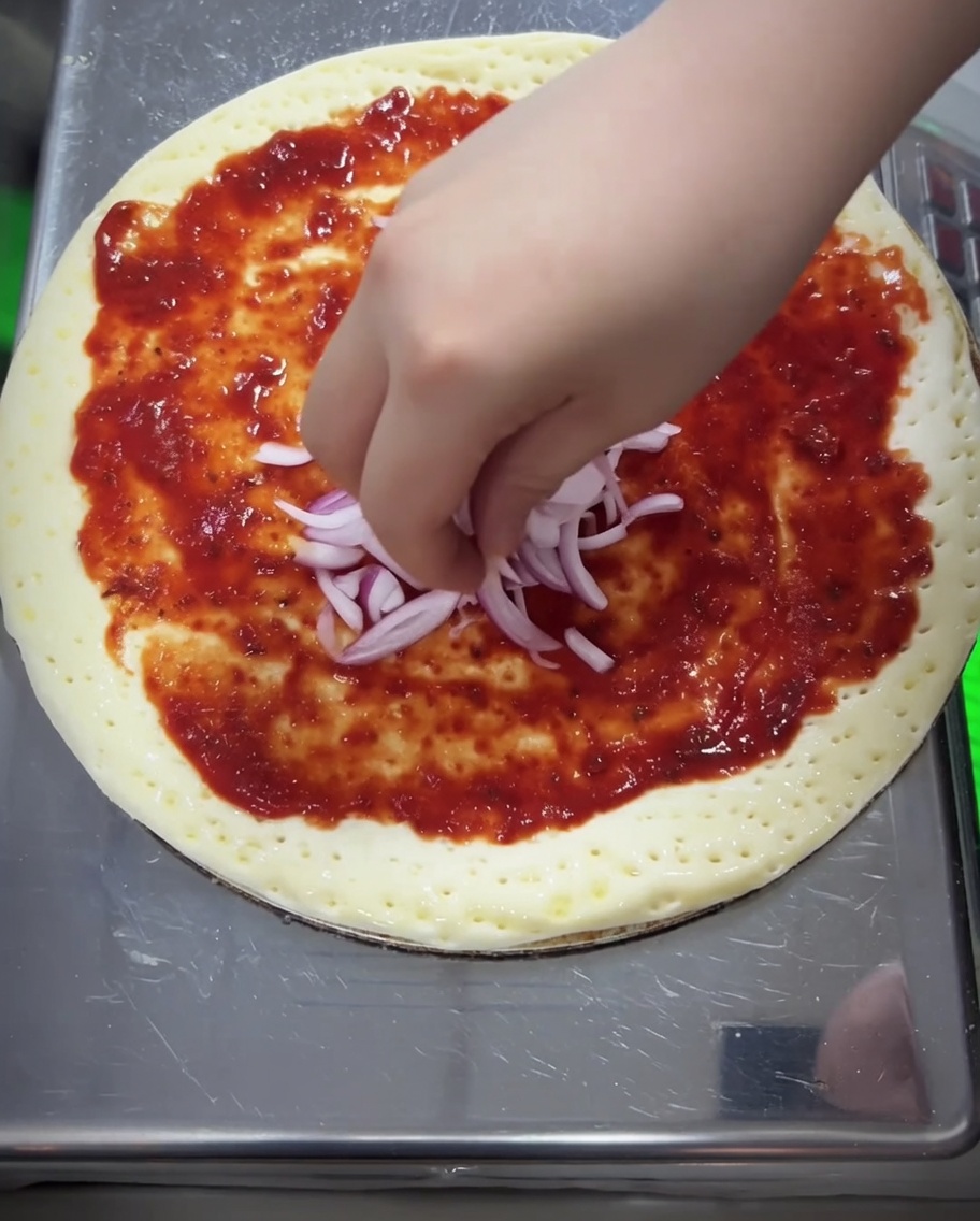 Czerwona cebula i starta mozzarella układane na sosie pomidorowym.