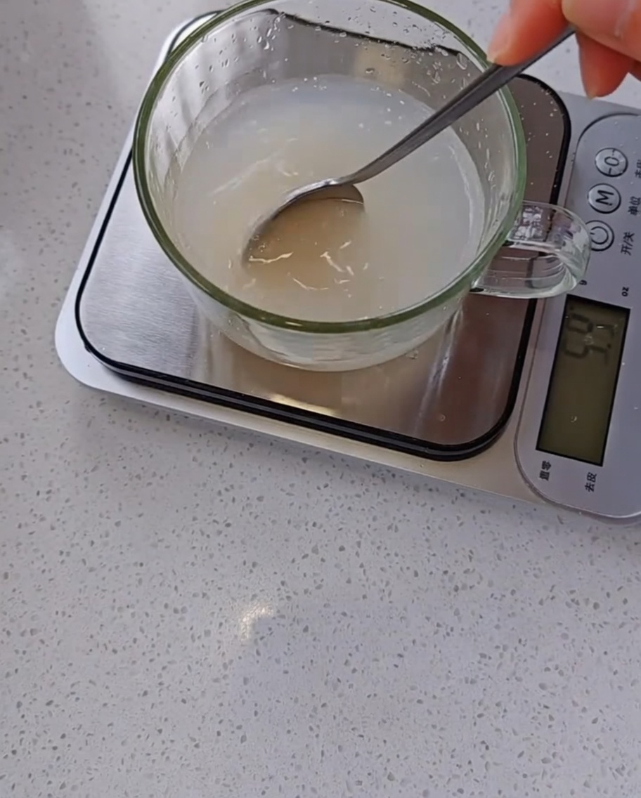 Dłoń mieszająca łyżką mętną mieszankę drożdży i wody w szklanej miarce ustawionej na wadze kuchennej.