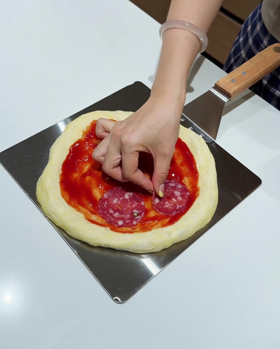 Dłoń kładąca okrągłe plastry salami na posmarowanym sosem spodzie z ciasta na metalowej łopacie.