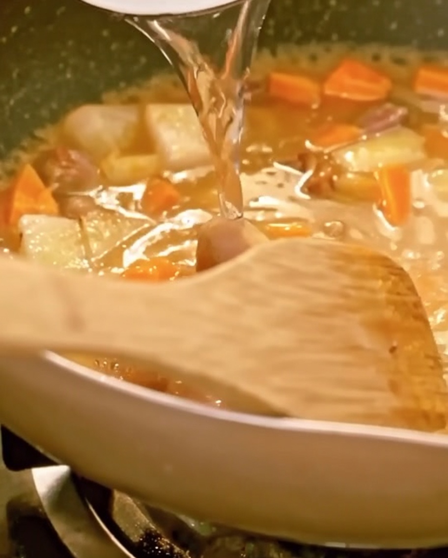 Woda wlewana na patelnię wypełnioną wołowiną, warzywami i topiącym się curry, widoczna drewniana łopatka.