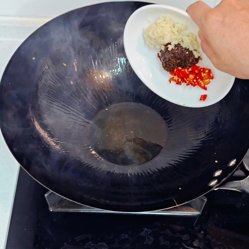 Dodawanie posiekanego czosnku, czerwonego chili i pasty sojowej do gorącego woka z małego, białego talerzyka.