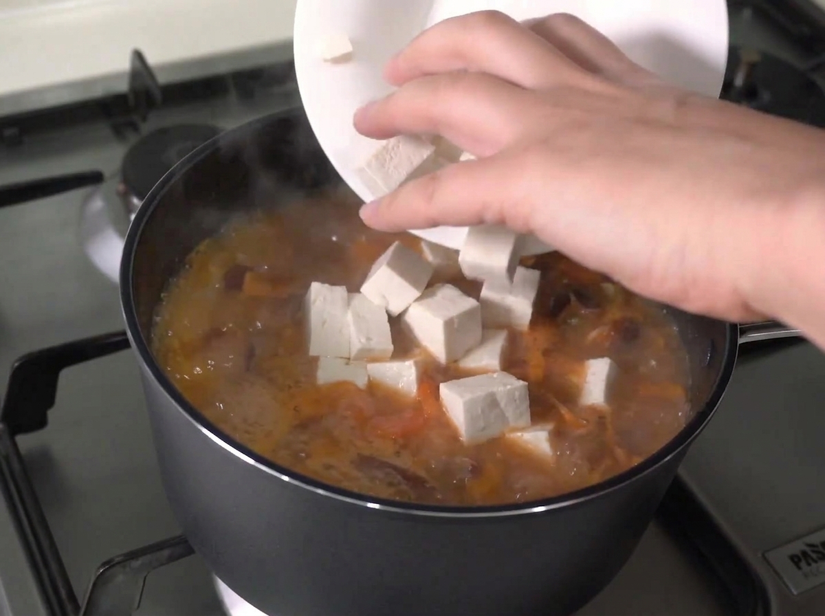 Świeże kostki tofu dodawane z białego talerza do aktywnie gotującego się gulaszu.