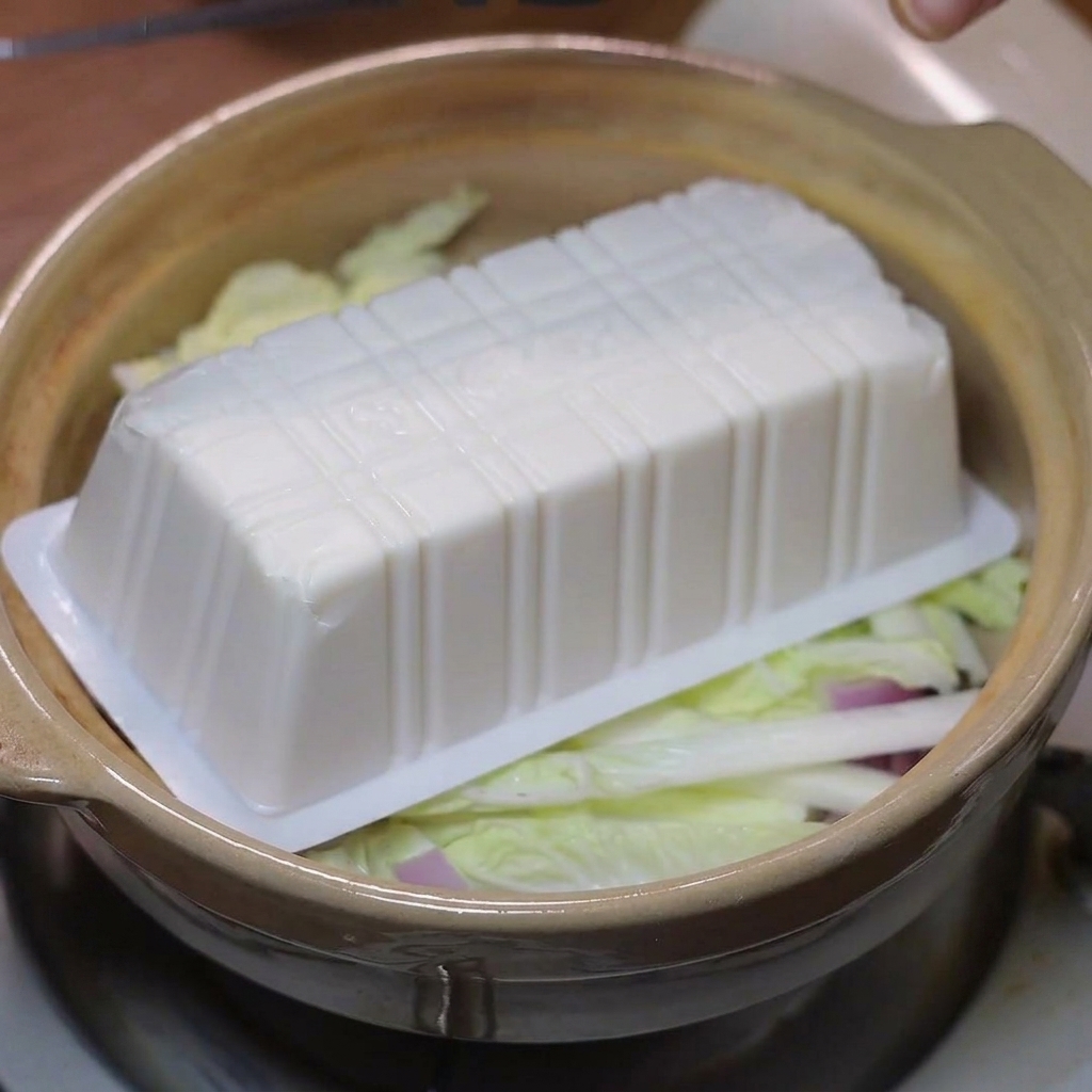 Kawałki miękkiego tofu ułożone na warstwie warzyw w glinianym garnku.