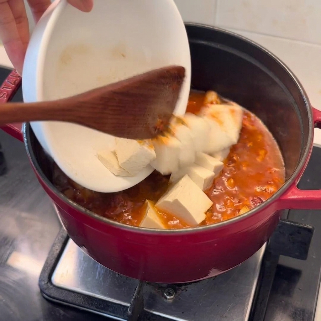 Świeże kostki tofu ostrożnie przekładane z białej miseczki do czerwonego garnka z gotującym się bulionem kimchi.