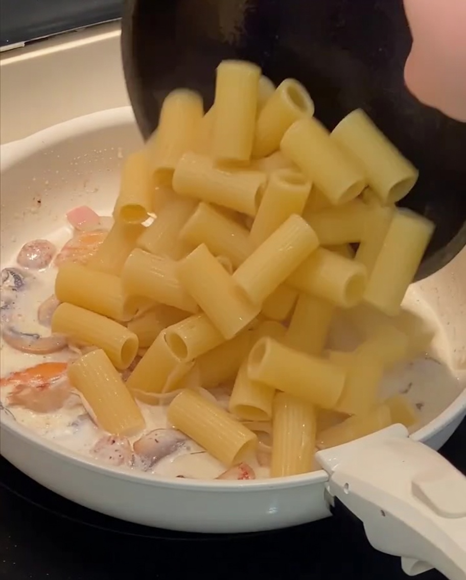 Ugotowany makaron rigatoni przekładany na patelnię z kremowym sosem.