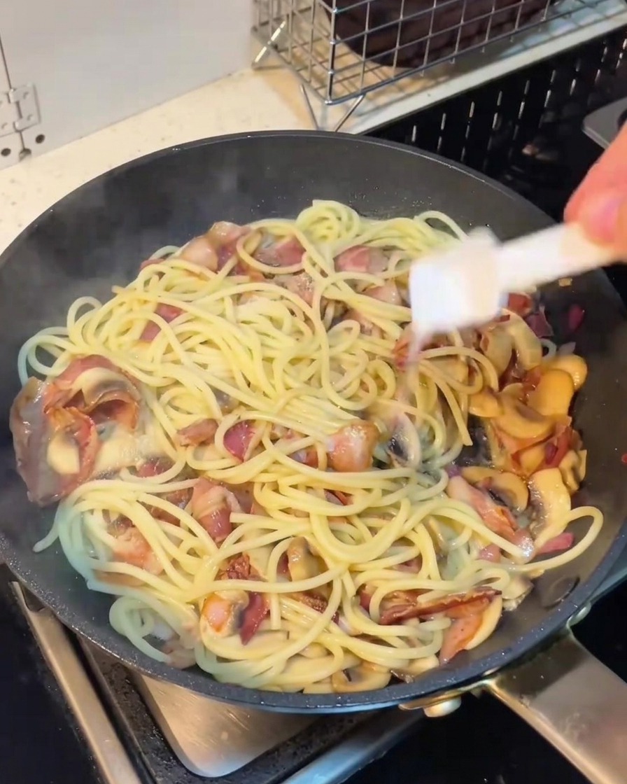 Ręka trzymająca przyprawnik nad patelnią ze spaghetti, boczkiem i pieczarkami.