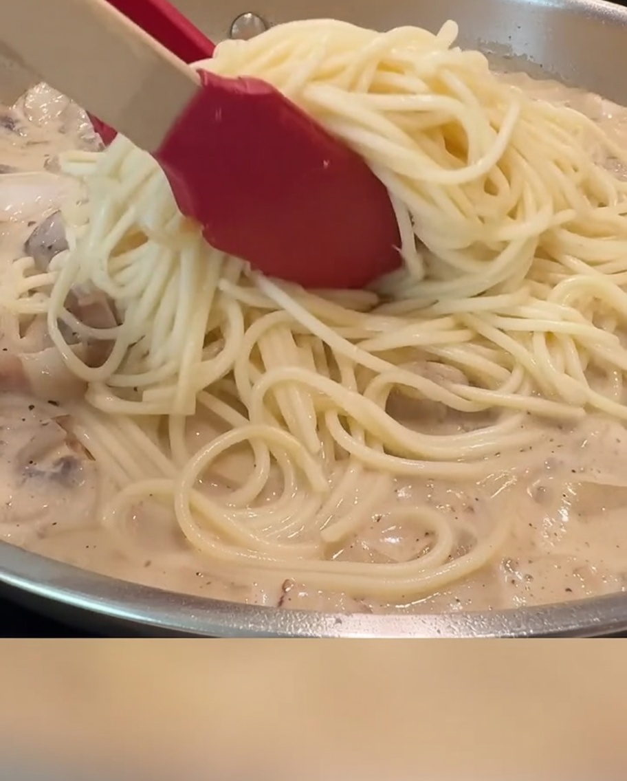 Spaghetti mieszane z sosem, boczkiem i pieczarkami na patelni za pomocą czerwonych szczypiec.