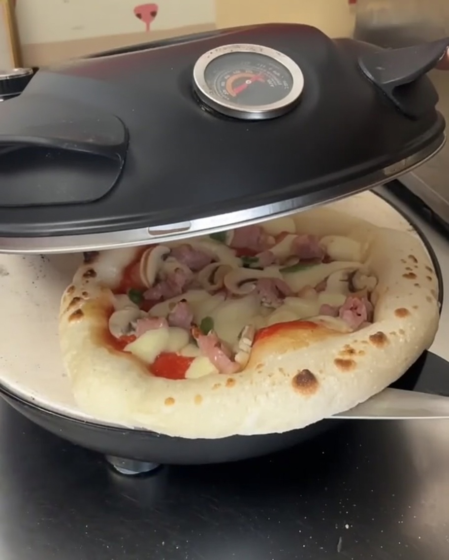 Podpieczona pizza z roztopionym serem wewnątrz otwartego pieca, obracana metalową łopatą.