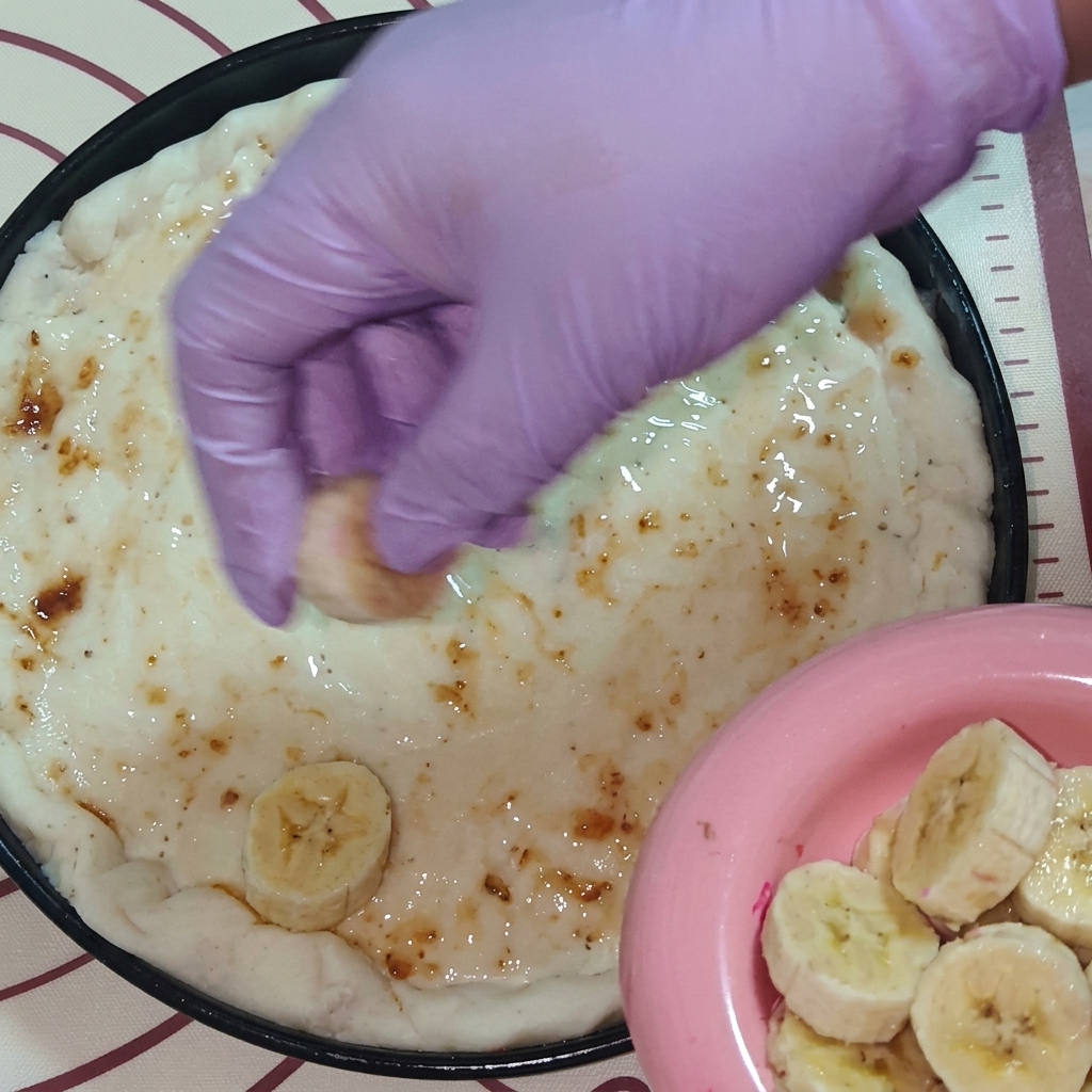 Dłoń w fioletowej rękawiczce układająca plastry świeżych owoców, w tym banana i smoczego owocu, na cieście pokrytym dżemem.