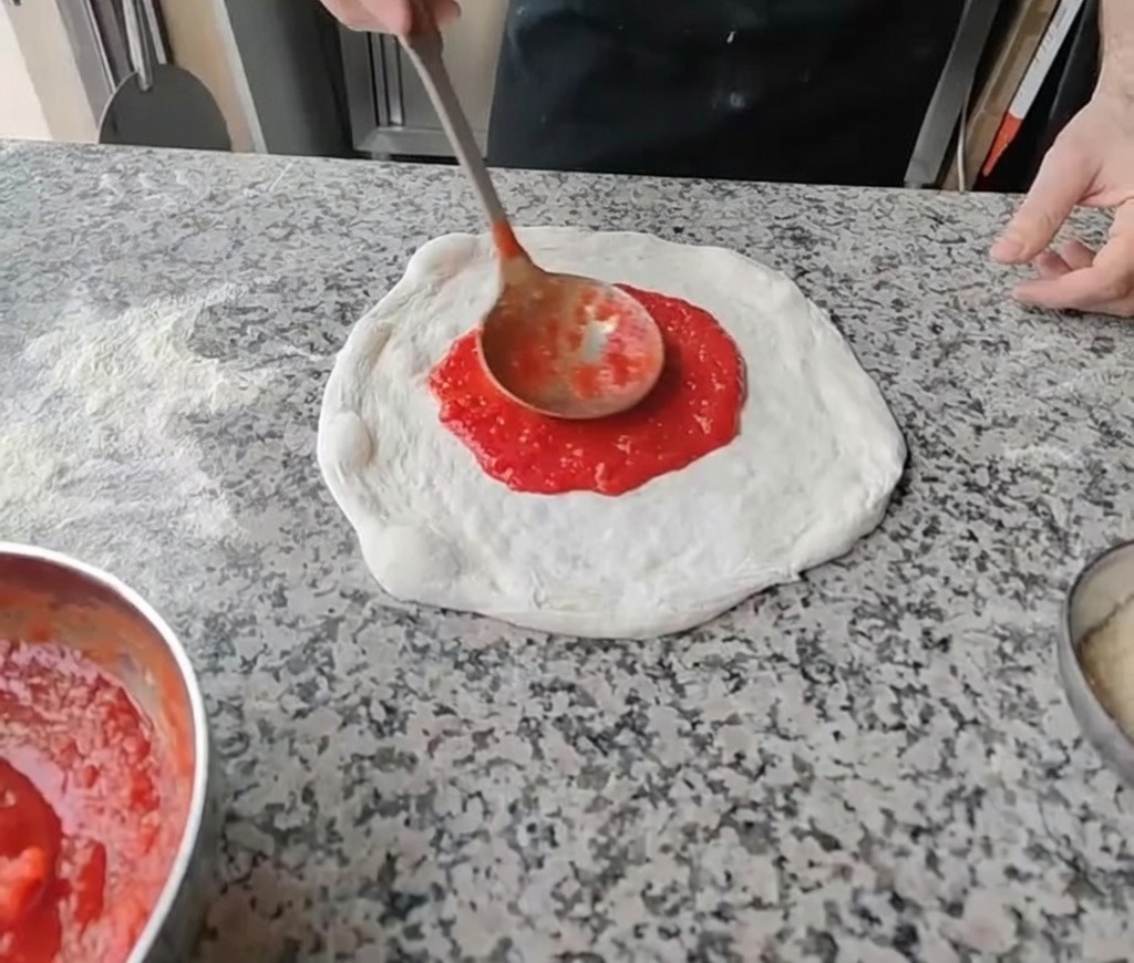 Rozsmarowywanie czerwonego sosu pomidorowego na środku rozciągniętego ciasta za pomocą chochli.