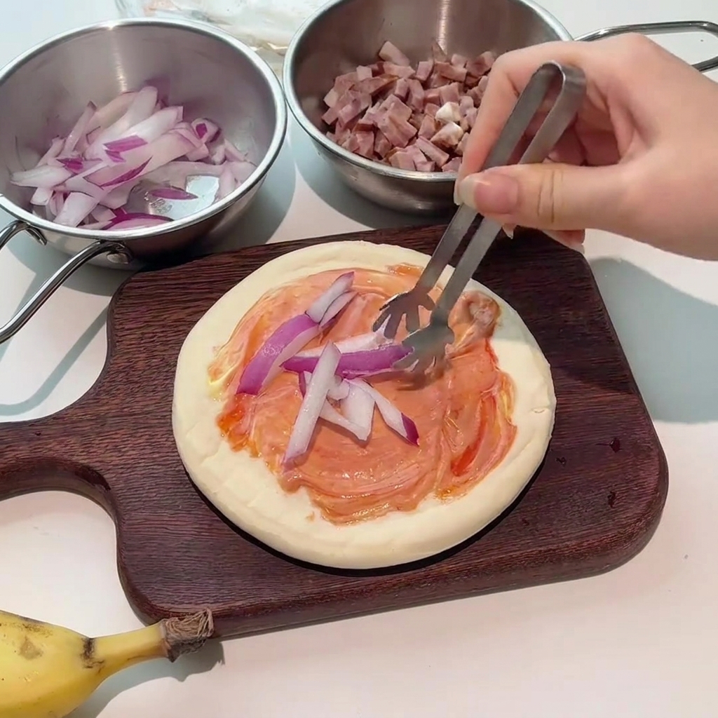 Szczypce układające świeże plasterki czerwonej cebuli na posmarowanym sosem spodzie do pizzy.