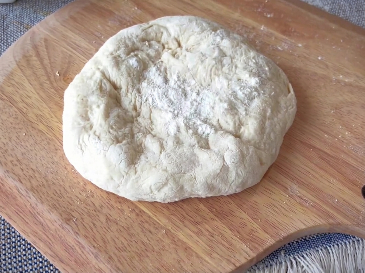 Okrągły kawałek ciasta na pizzę rozciągany na drewnianej powierzchni z wyraźnie zaznaczonym grubym brzegiem.