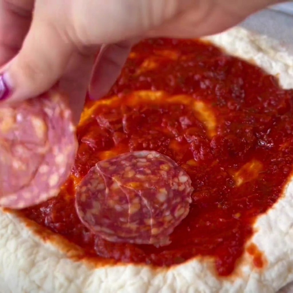 Plasterki salami ostrożnie układane na posmarowanym sosem środku pizzy.