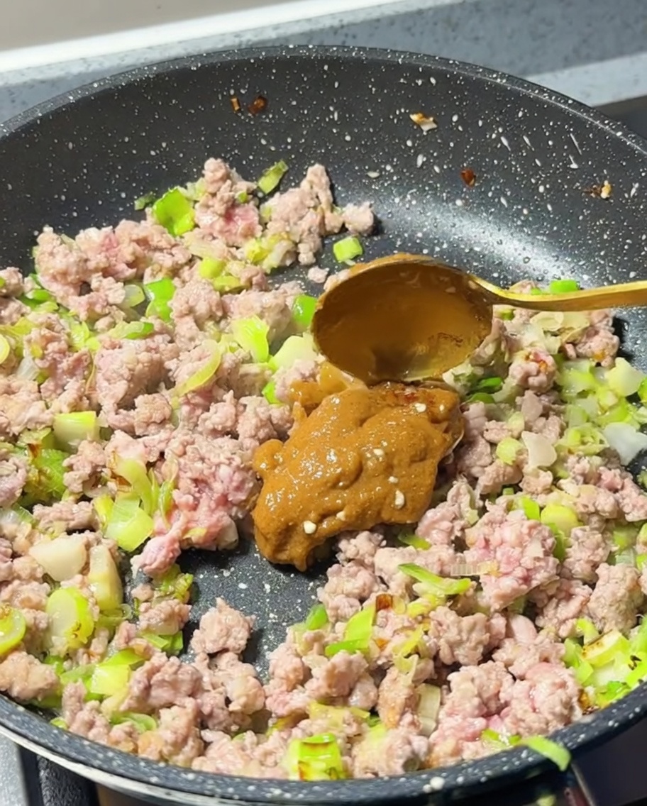 Łyżka jasnobrązowej pasty miso dodawana do patelni z usmażonym mięsem.