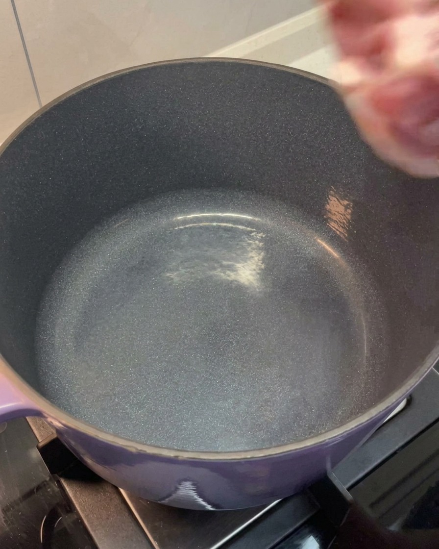 Opłukany boczek i kości z łopatki wkładane do dużego fioletowego garnka.