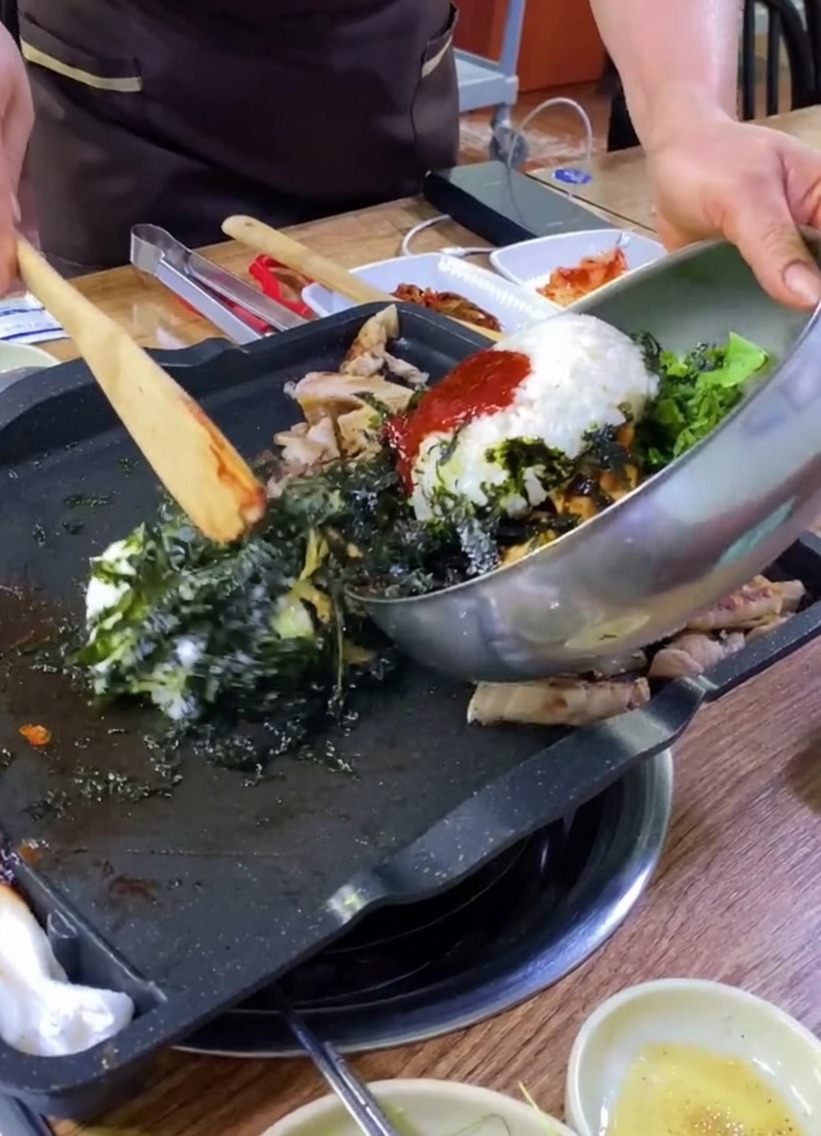 Ryż, warzywa i wodorosty przesypywane z metalowej miski na grill z boczkiem.