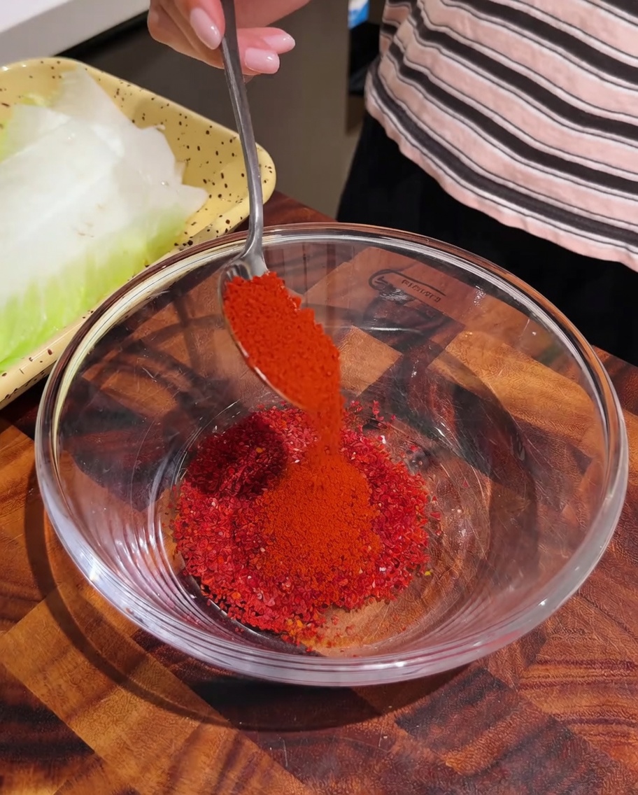 Czerwone chili w proszku wsypywane łyżką do szklanej miski zawierającej inne przyprawy.