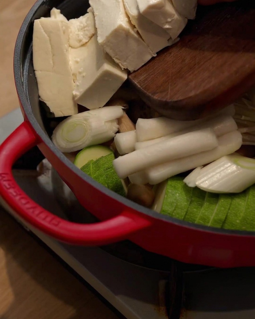 Bloki tofu zsuwane z drewnianej deski do garnka zawierającego inne składniki.