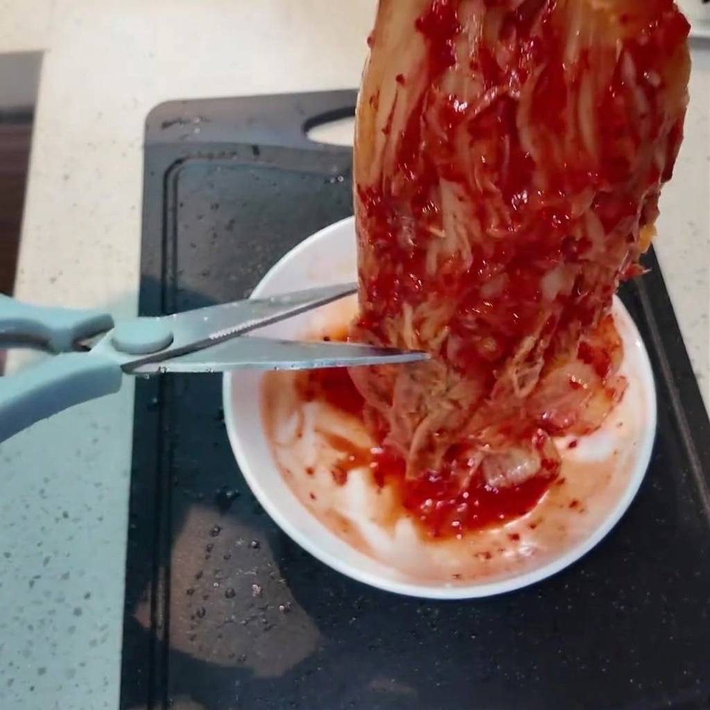 Kawałek jaskrawo czerwonego kimchi cięty na mniejsze kawałki za pomocą nożyczek kuchennych nad małym białym talerzem.