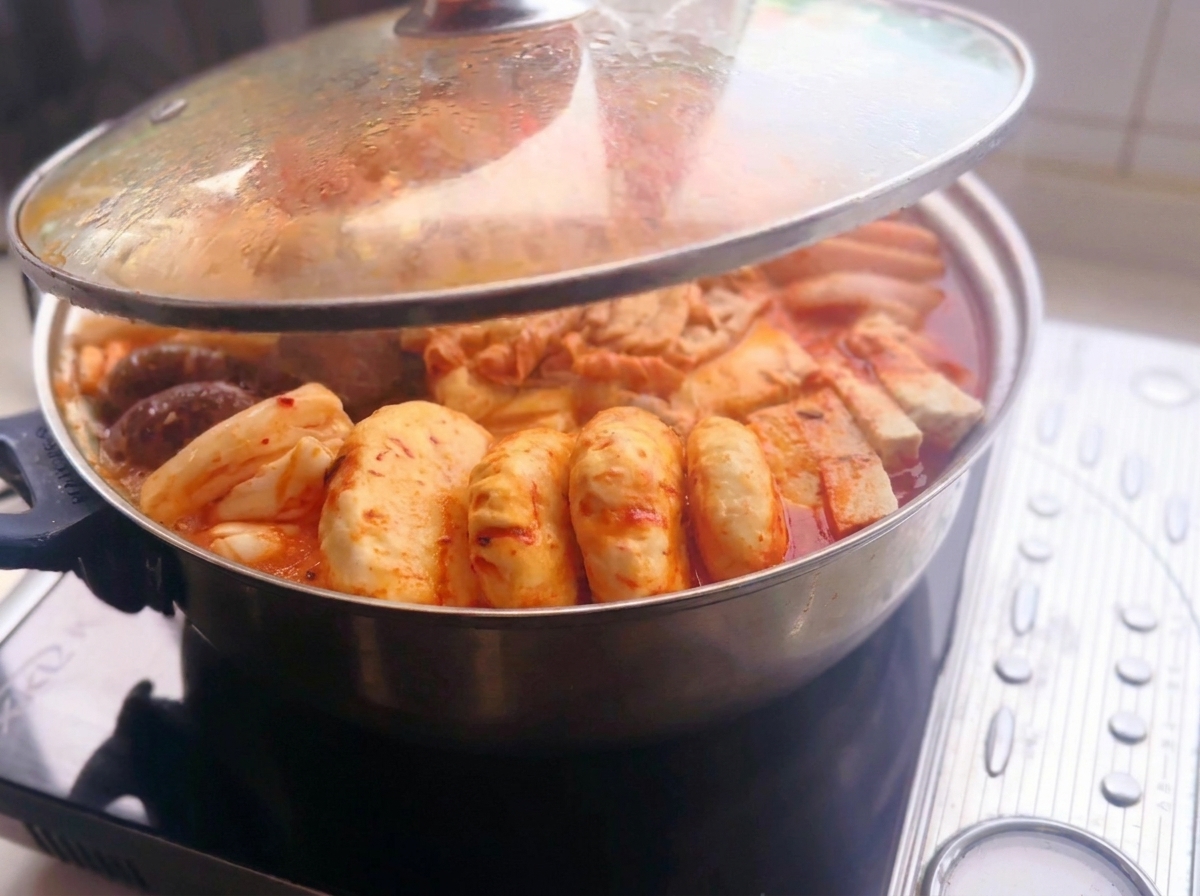 Szklana pokrywka uniesiona nad metalowym garnkiem wypełnionym ułożonymi składnikami, takimi jak pokrojone mięso, tofu, grzyby, ryżowe ciasteczka i pierożki w pikantnym czerwonym bulionie na kuchence indukcyjnej.