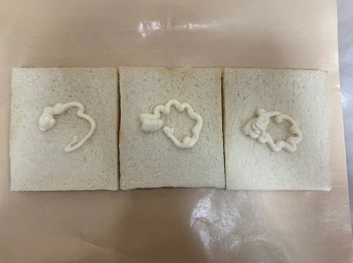 Trzy kromki białego chleba bez skórki ułożone obok siebie na brązowym papierze do pieczenia, każda polana esencjonalną linią białego sosu sałatkowego.