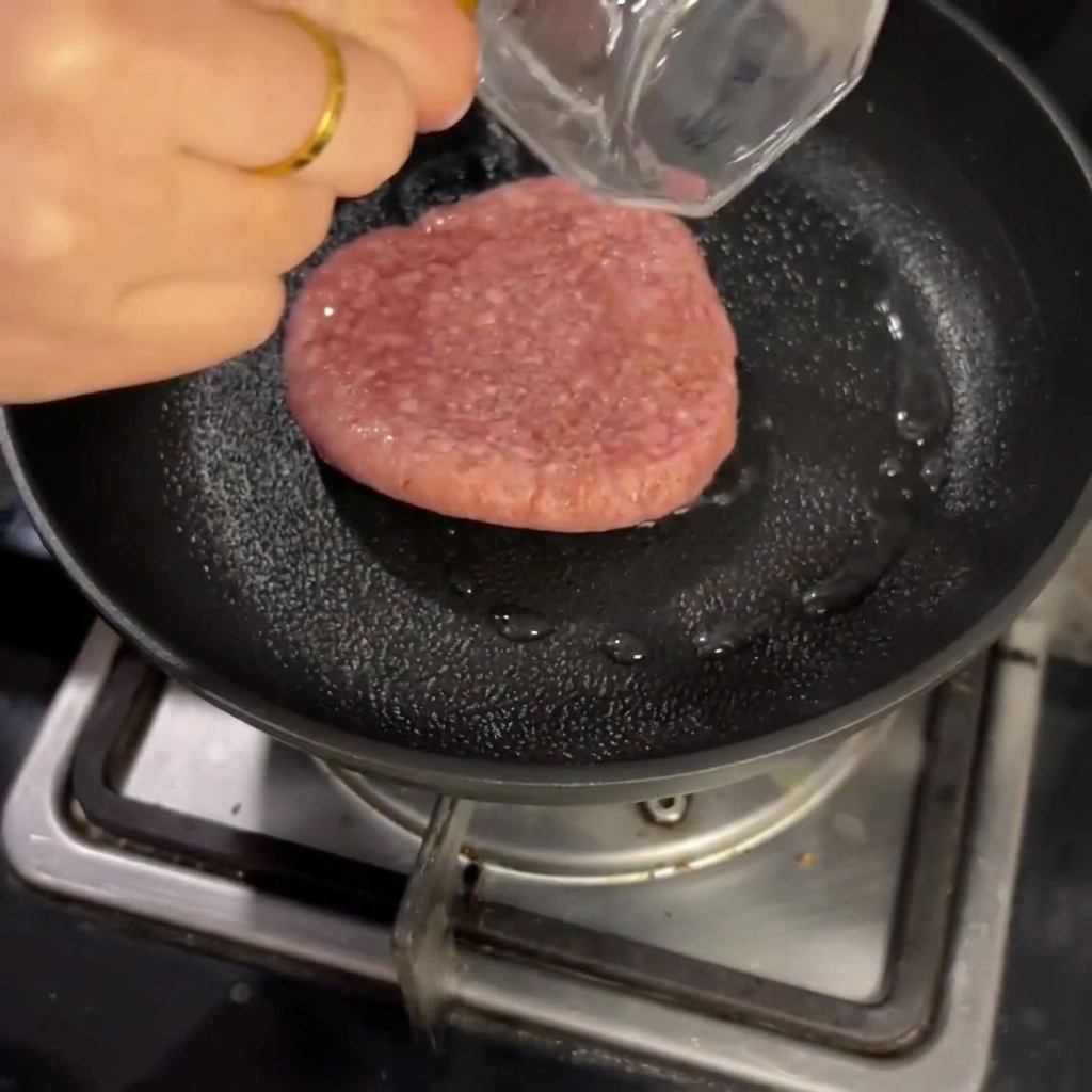 Surowy kotlet wołowy smażony na patelni z niewielką ilością płynu, pod przykryciem.