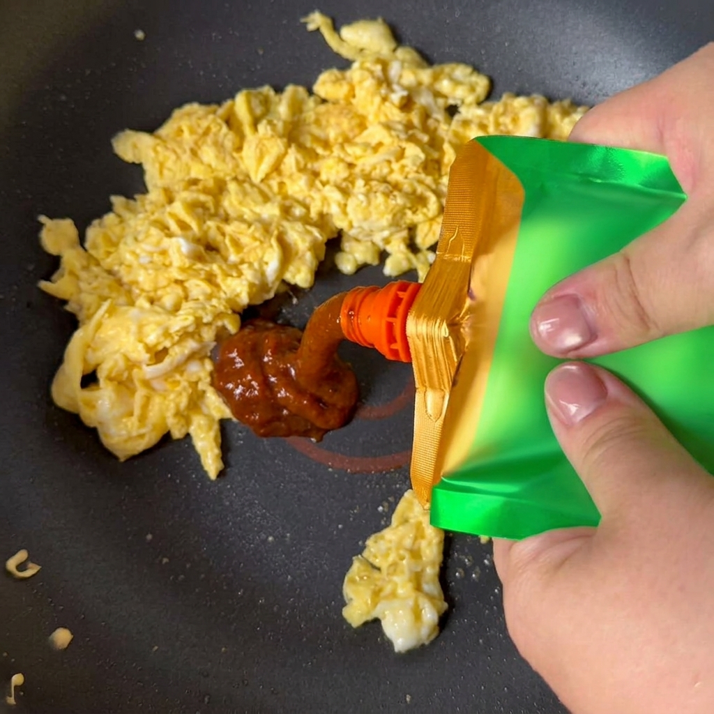 Żółta pasta sojowa wyciskana z zielonej saszetki na jajecznicę na patelni.