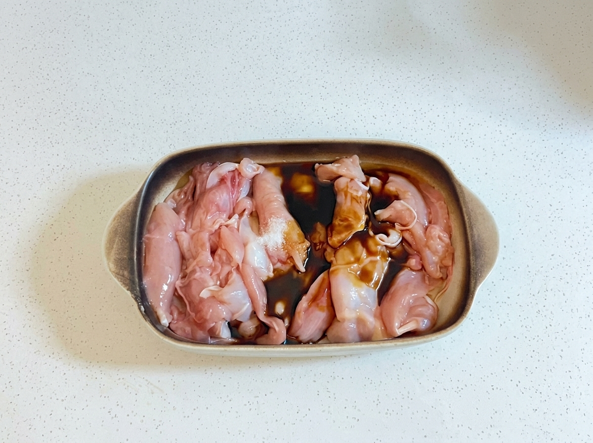 Ciemne płyny kuchenne i olej wlewane na surowe kawałki kurczaka w naczyniu do zapiekania.
