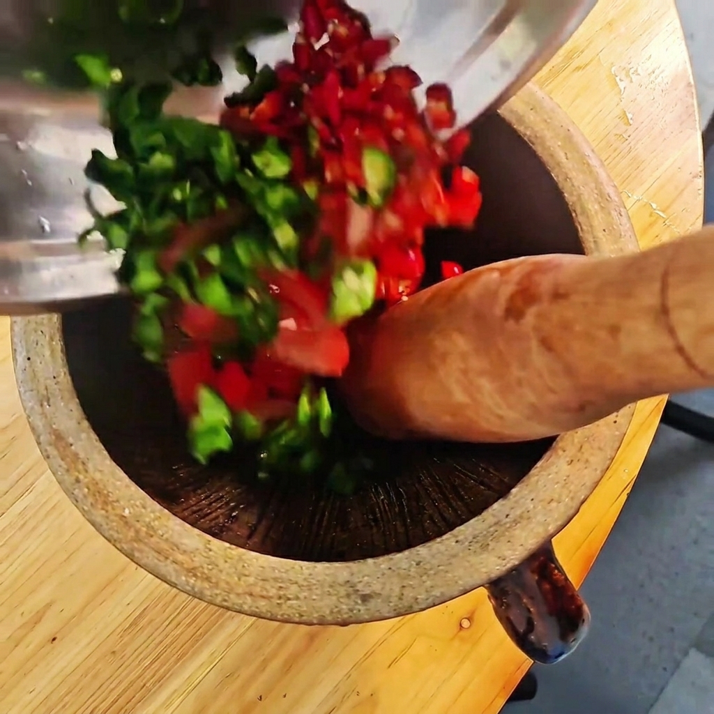 Posiekane zielone i czerwone papryczki chili oraz pomidory ucierane w drewnianym moździerzu.
