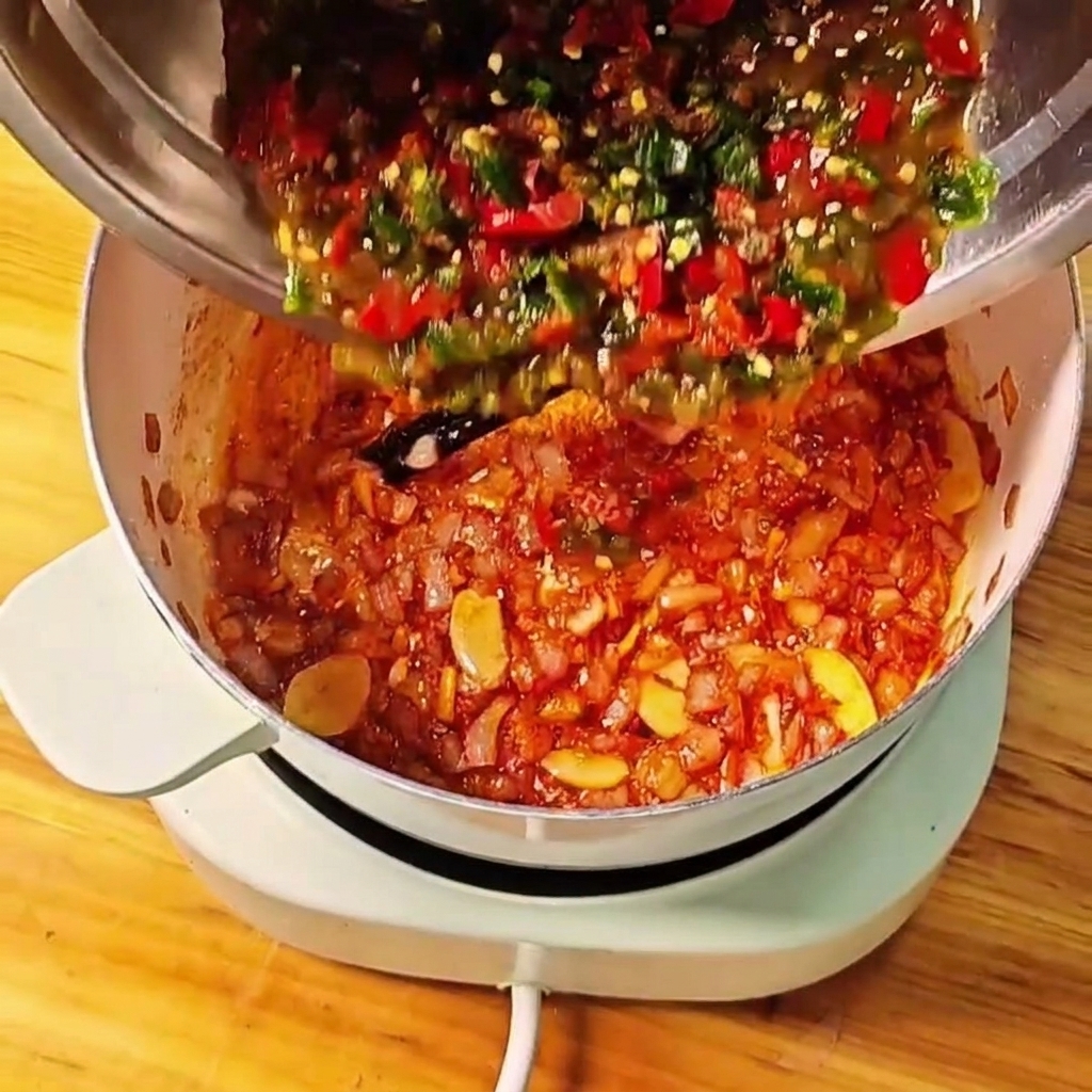 Rozgniecione pomidory z papryczkami chili wlewane do garnka z aromatyczną bazą.