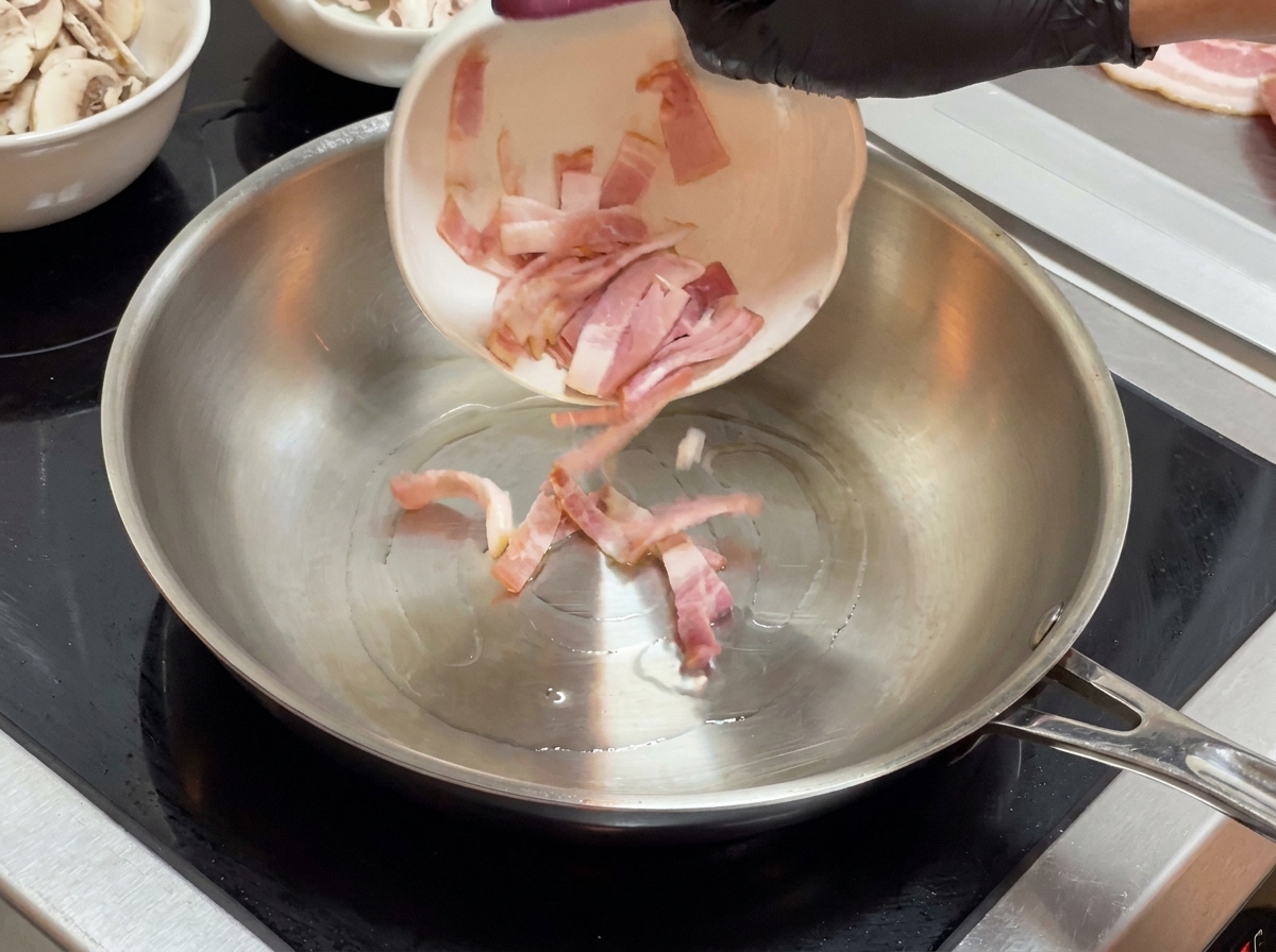 Kawałki boczku wsypywane z małej białej miski na patelnię ze stali nierdzewnej.