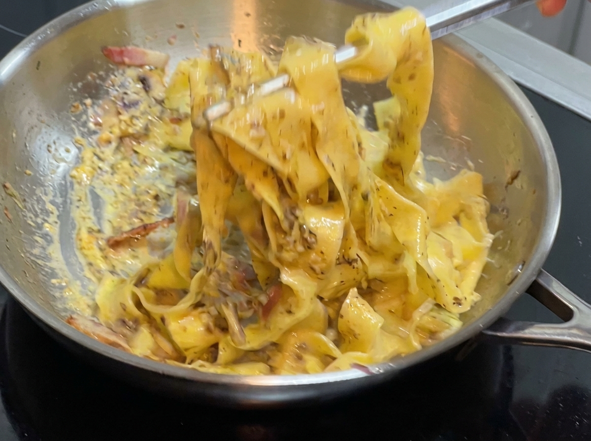 Szczypcy mieszające szeroki makaron i sos na patelni ze stali nierdzewnej na kuchence.