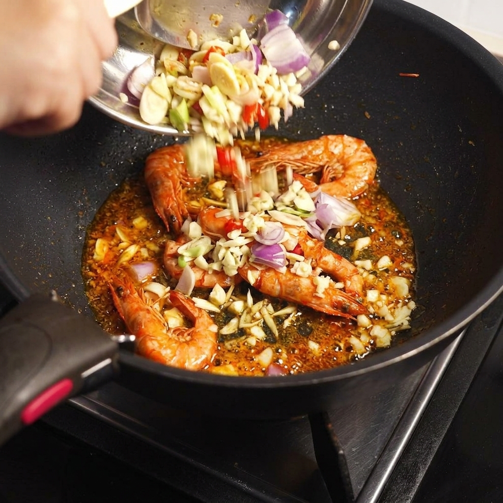 Posiekane składniki aromatyczne i pasta Tom Yum dodawane z miski na patelnię ze smażonymi pancerzami krewetek.