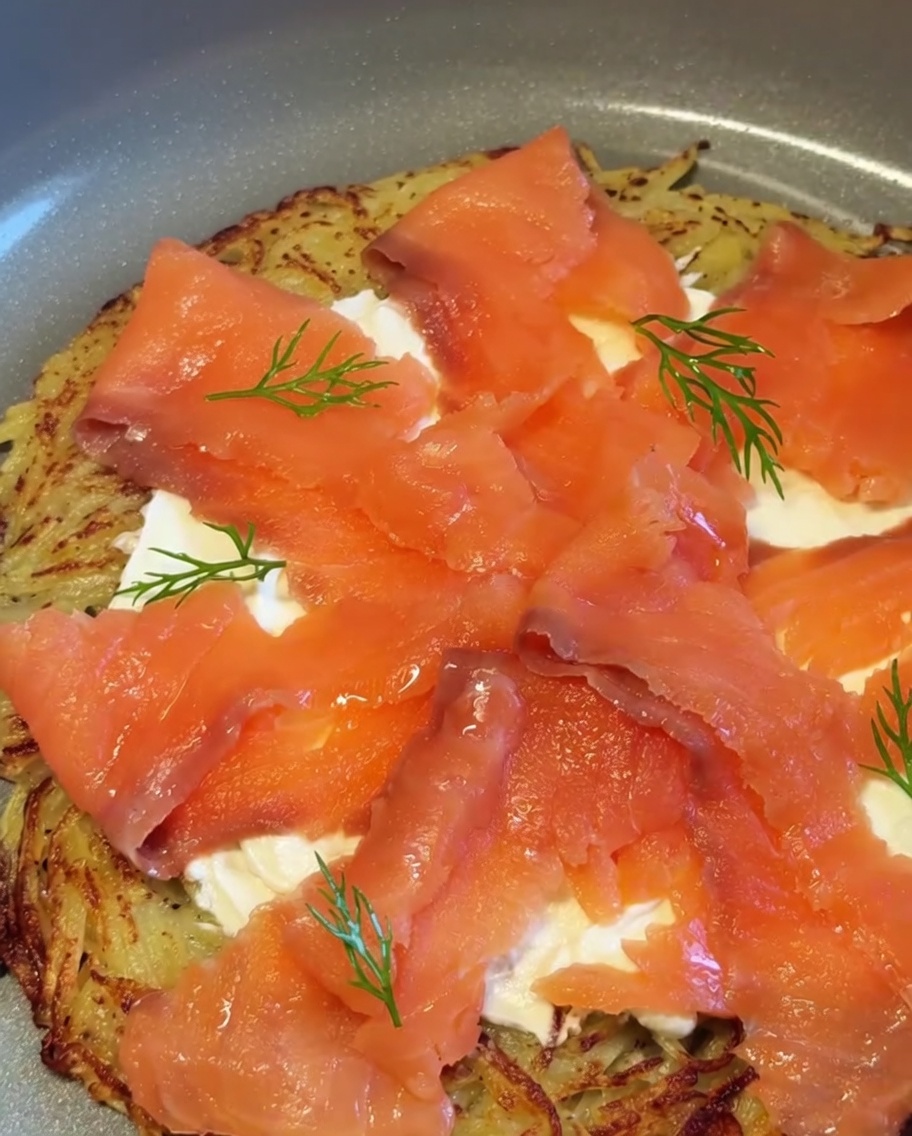Gotowa pizza ziemniaczana z wędzonym łososiem udekorowana gałązkami świeżego koperku i kawałkami czerwonej cebuli.